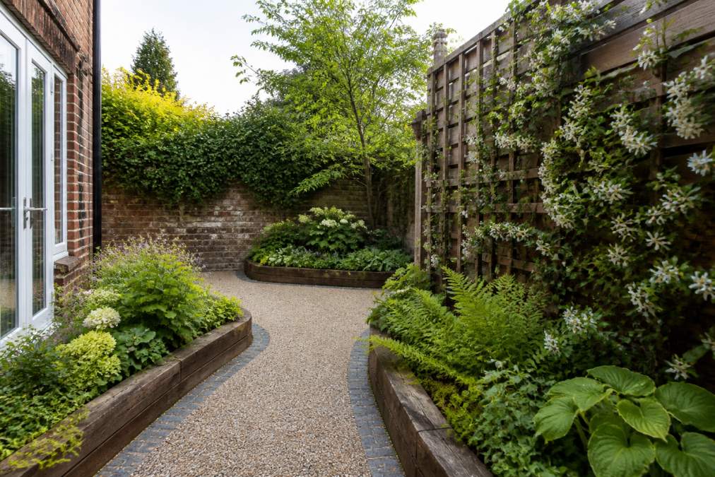 A small courtyard garden in a UK home with a curved resin bound path, raised timber beds, and a vertical trellis of climbing plants, photographed in soft afternoon light