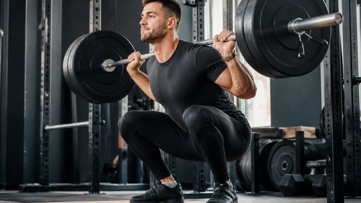 Male weightlifter performing a deep barbell back squat in the gym, wearing black muscle-fit training trousers that show clear freedom of movement through the quads and hips, with a loaded barbell across his shoulders