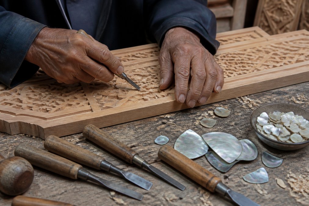 Close-up of master artisan's weathered hands carving intricate geometric patterns into pale cedarwood using traditional hand tools, with mother of pearl shell pieces and carving implements arranged on a wooden workbench