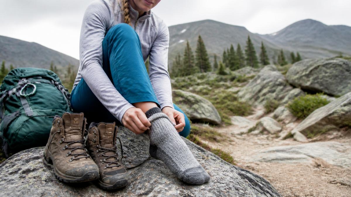 Close-up of a hiker sitting on a trailside rock, pulling an alpaca crew sock over their foot beside a worn hiking boot, with a daypack and rocky trail visible behind them