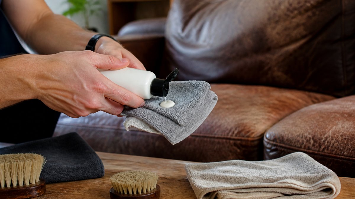 Close-up of a person's hands applying leather cleaner to a soft microfibre cloth beside a worn brown leather sofa in a warm living room