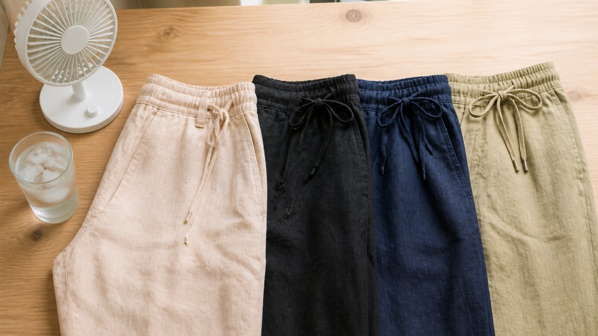 Four pairs of linen trousers in neutral colors with a desk fan and iced water on a wooden surface