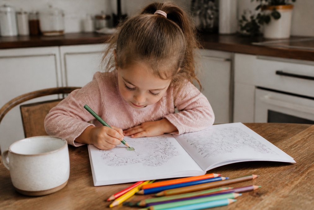 A young child sitting at a wooden kitchen table, deeply focused on a coloring page in an open activity book, a few colored pencils scattered nearby in soft natural light