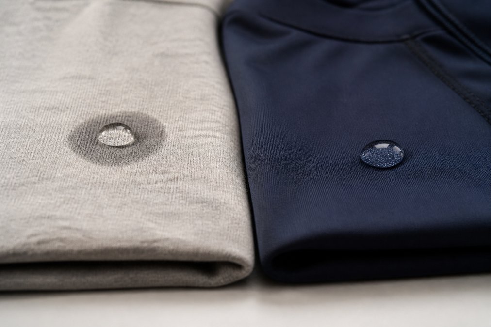 A close-up photograph of two folded gym shirts side by side on a clean surface, one made of cotton and one made of a smooth synthetic performance fabric, with a water droplet resting on each fabric surface to show how differently they absorb moisture