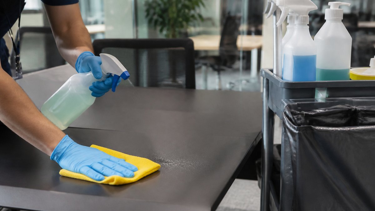 Cleaner in gloves wiping an office desk with a spray bottle and cloth near a janitorial supply trolley