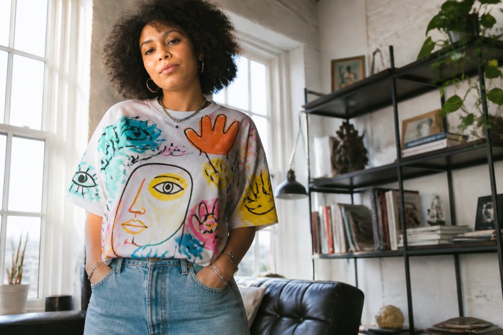 A person wearing a colorful artistic graphic t-shirt with bold hand-drawn style prints, standing confidently in a bright casual loft setting with natural window light