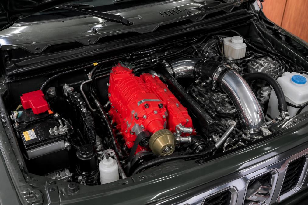 Close-up of a supercharger kit mounted on a Suzuki engine with visible polished piping, intake manifold, and belt-drive system in a well-lit workshop