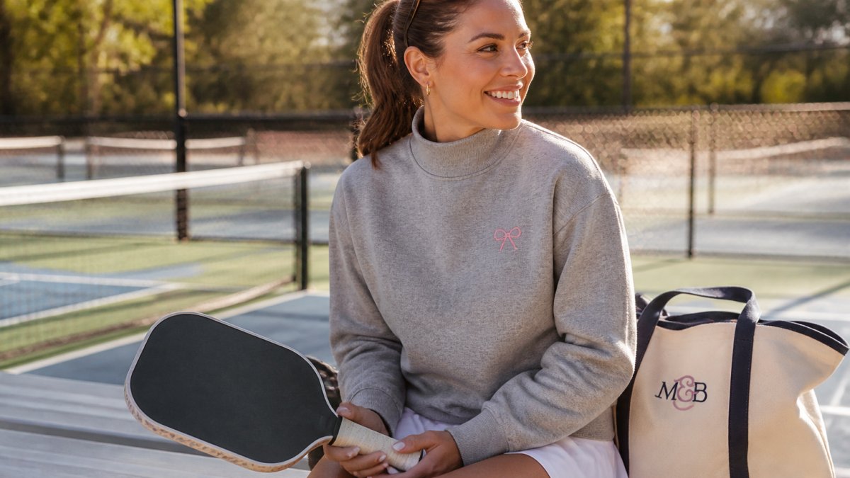 A woman in a heather gray mock neck sweatshirt with pink embroidered detail, holding a pickleball paddle and smiling on a sunny court, a canvas bag resting nearby