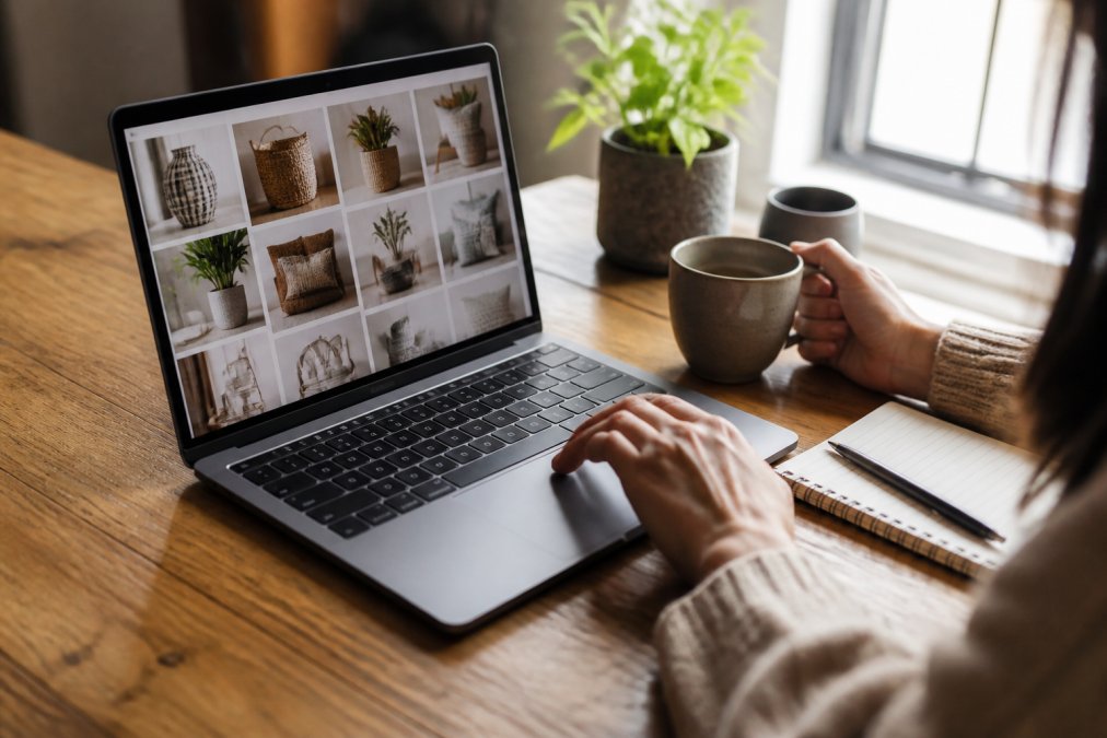 Close-up of a laptop screen displaying an online marketplace with home decor items, a cup of coffee and notepad nearby on a wooden desk, natural afternoon light streaming through a window