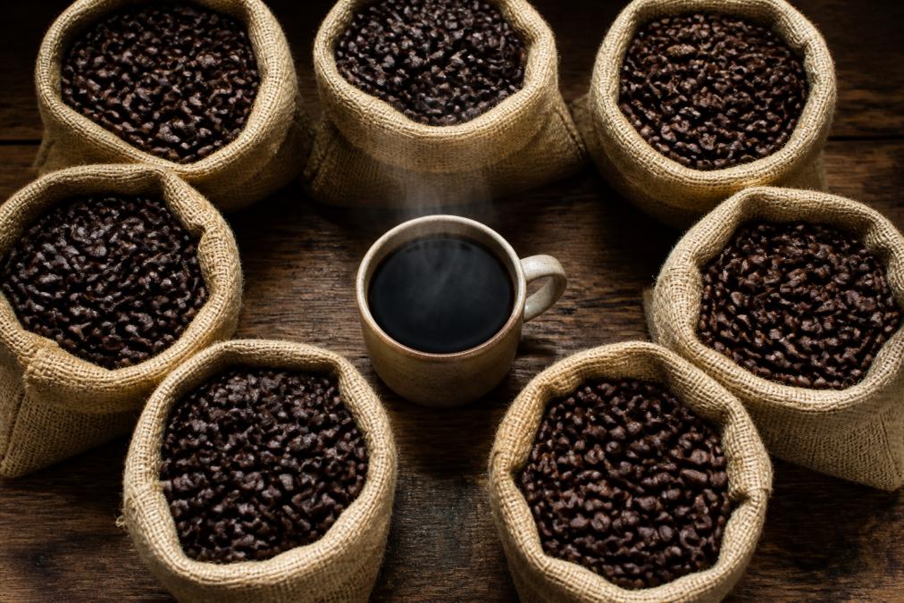 An overhead shot of several open burlap coffee bags filled with dark roasted whole beans, each labeled, on a rustic wooden table with a steaming ceramic cup of black coffee in the center