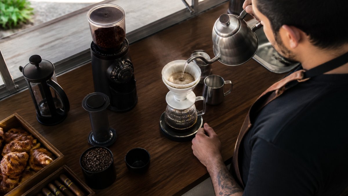 Overhead shot of a home coffee brewing station with whole beans, a grinder, pour-over dripper, French press, AeroPress, and a kettle arranged on a wooden counter with natural morning light