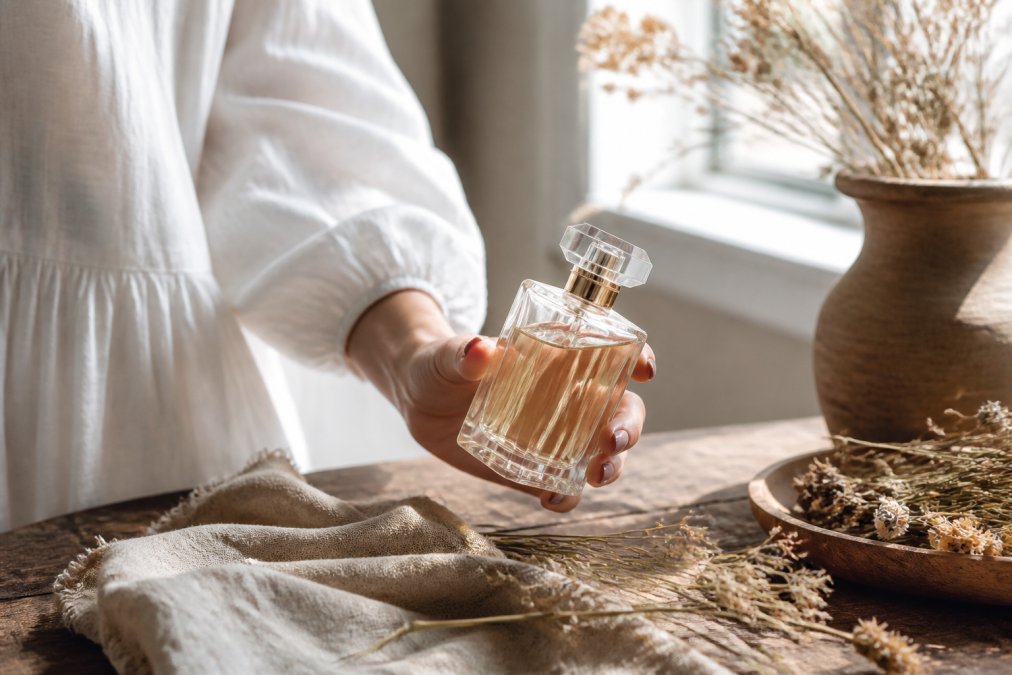 A woman's hand holding an elegant glass perfume bottle with soft natural light filtering through a window, dried flowers and a linen cloth on a wooden table nearby