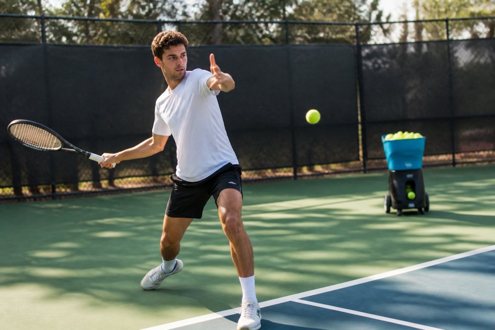 A tennis player in a dynamic ready position executing a forehand from the baseline during a practice session, with a ball machine visible in the background