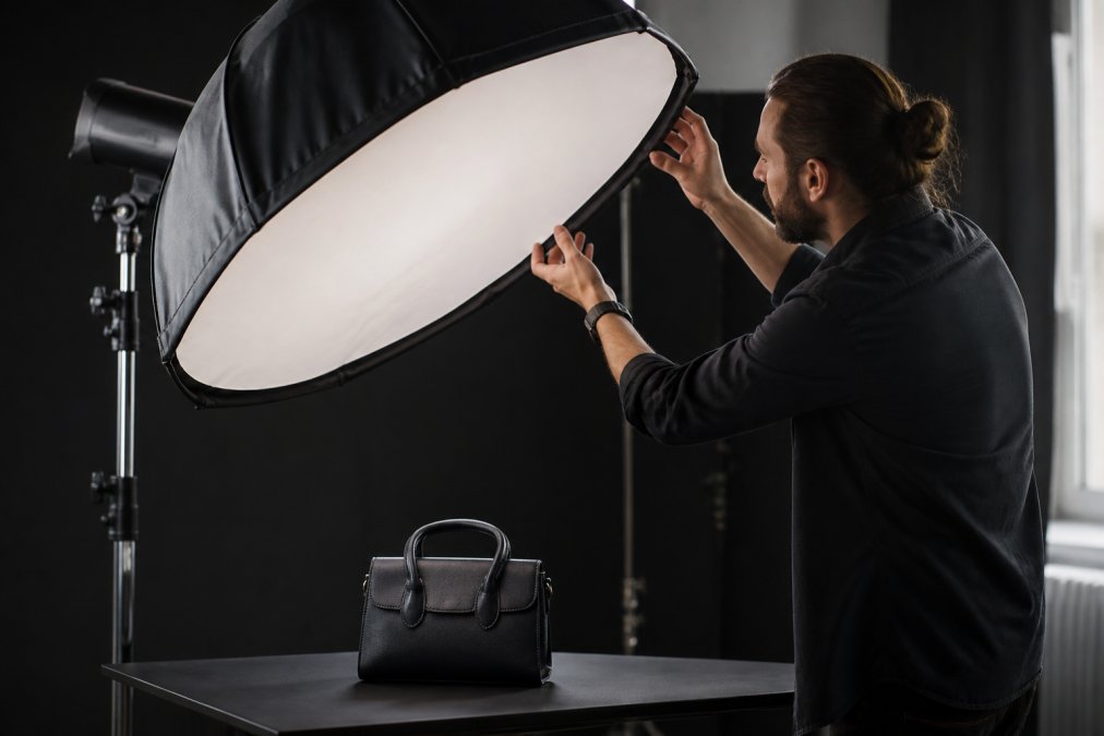 A professional photographer adjusting a softbox and diffuser to light a product on a table in a studio setting, with a dark backdrop to emphasize the lighting setup