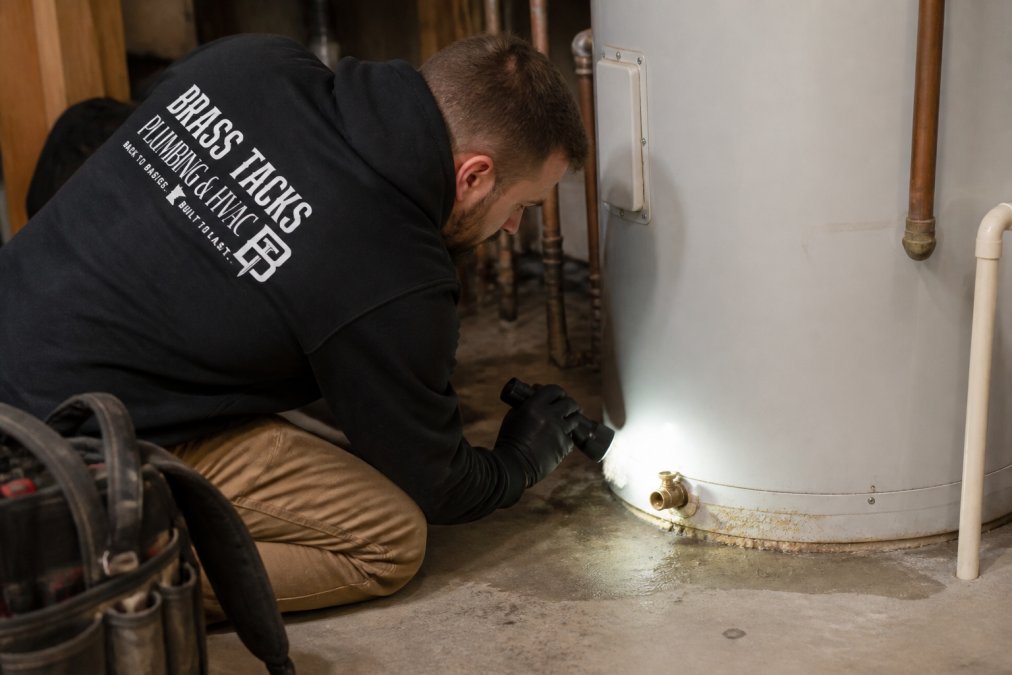 A licensed plumber in a residential basement inspecting the bottom of a Bradford White water heater tank, with a flashlight illuminating visible sediment buildup near the drain valve