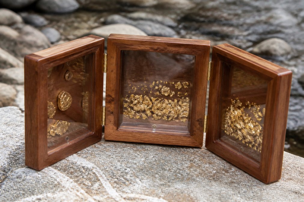 Close-up of raw gold flakes and small nuggets displayed in a handcrafted wooden tri-fold case with clear acrylic windows, warm lighting revealing the natural luster and texture of the placer gold against rich wood grain