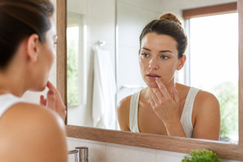 Close-up of a person touching their lip with a concerned expression, standing in a bright bathroom mirror setting, natural light