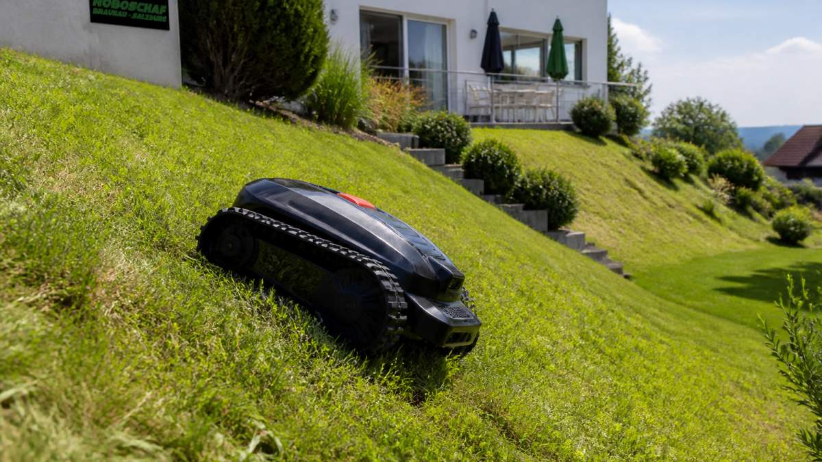 Ein moderner Mähroboter mit Raupenfahrwerk mäht bei sonnigem Wetter eine steile, grasbewachsene Böschung, im Hintergrund ein Wohnhaus mit Garten