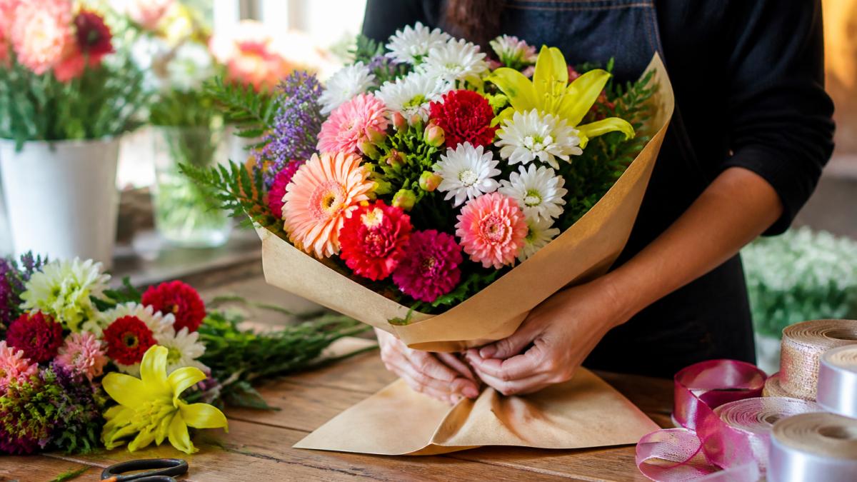 Florist wrapping a colorful birthday flower delivery bouquet in kraft paper at a sunlit wooden workbench