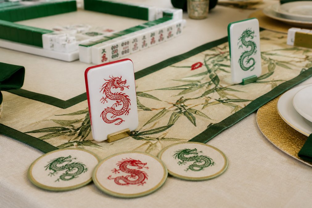Close-up of an elegantly set mahjong game table with red and green dragon-themed tile-shaped place cards, a coordinating fabric table runner with bamboo motifs, and decorative coasters arranged on a cream-colored tablecloth
