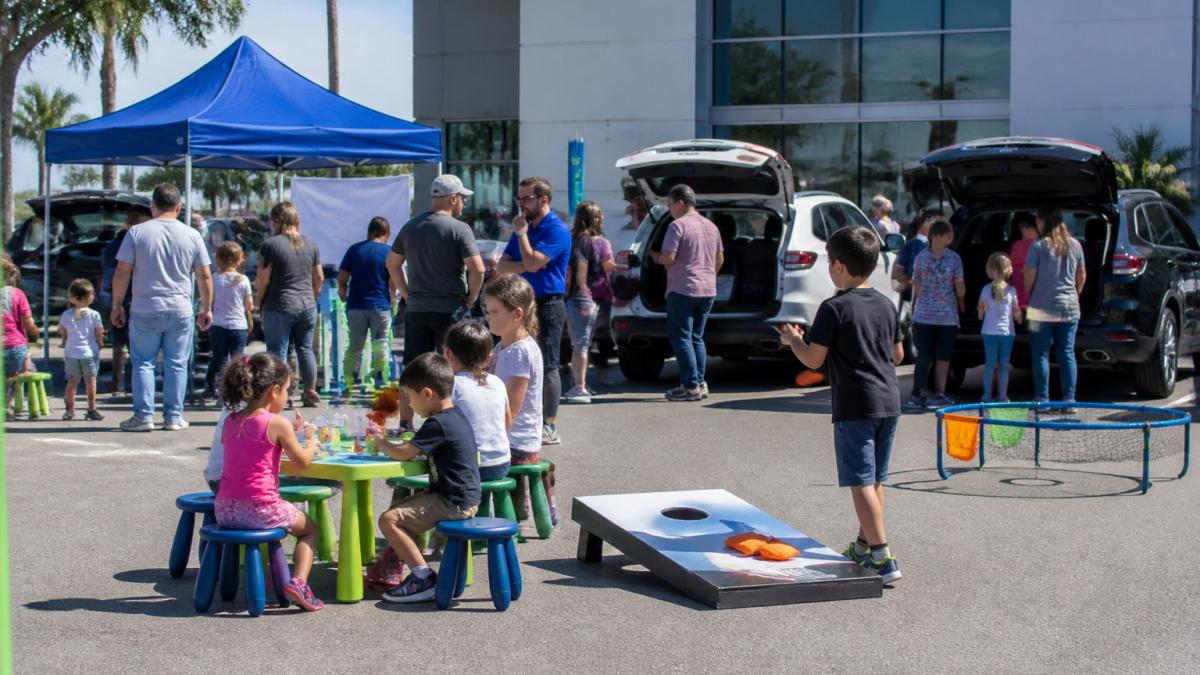 Lively car dealership event with families browsing vehicles, children enjoying outdoor activities near branded tents and display cars, staff engaging with visitors, sunny afternoon lighting