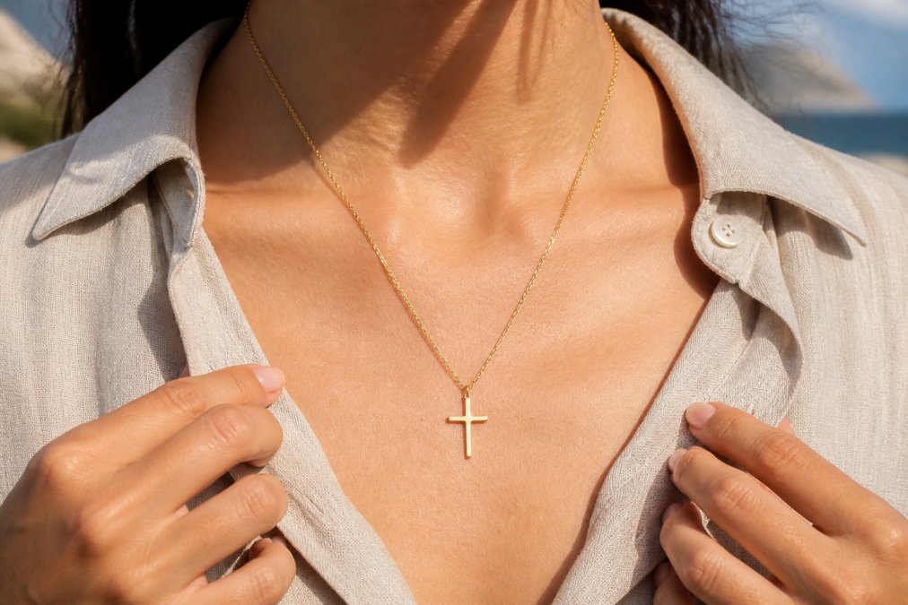 Person wearing a gold-plated cross necklace over a casual linen shirt in natural daylight, showing the necklace against skin with visible detail of the pendant's finish