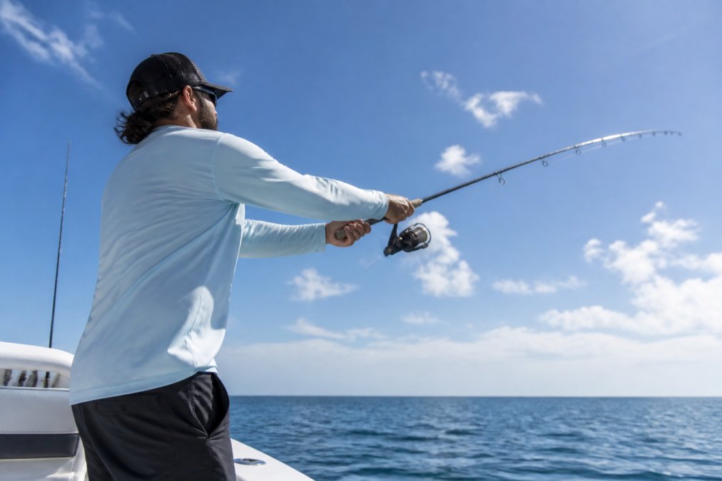Angler wearing a long-sleeve performance fishing shirt standing on a boat in bright sunlight, casting a fishing rod with ocean and sky in background