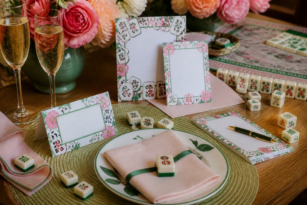 Colorful mahjong-themed table setup with printed place cards, invitation suite, and coordinating stationery spread across wooden table with mahjong tiles and champagne glasses visible