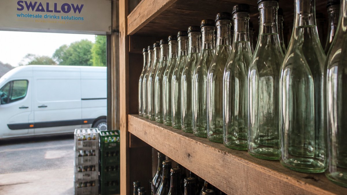 Lightweight glass bottles on a pub cellar shelf with crates and a delivery van visible through the open door