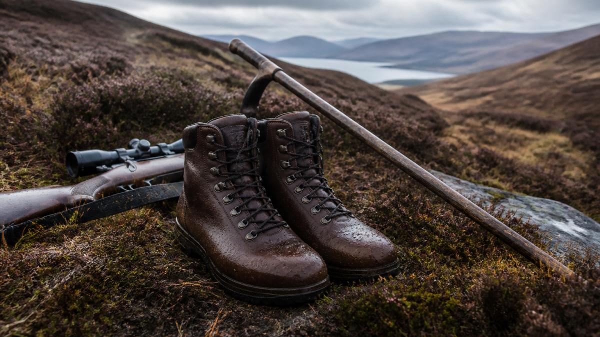 A pair of brown leather laced country boots standing on wet heather moorland, with a rifle and walking stick resting nearby, soft overcast Scottish light
