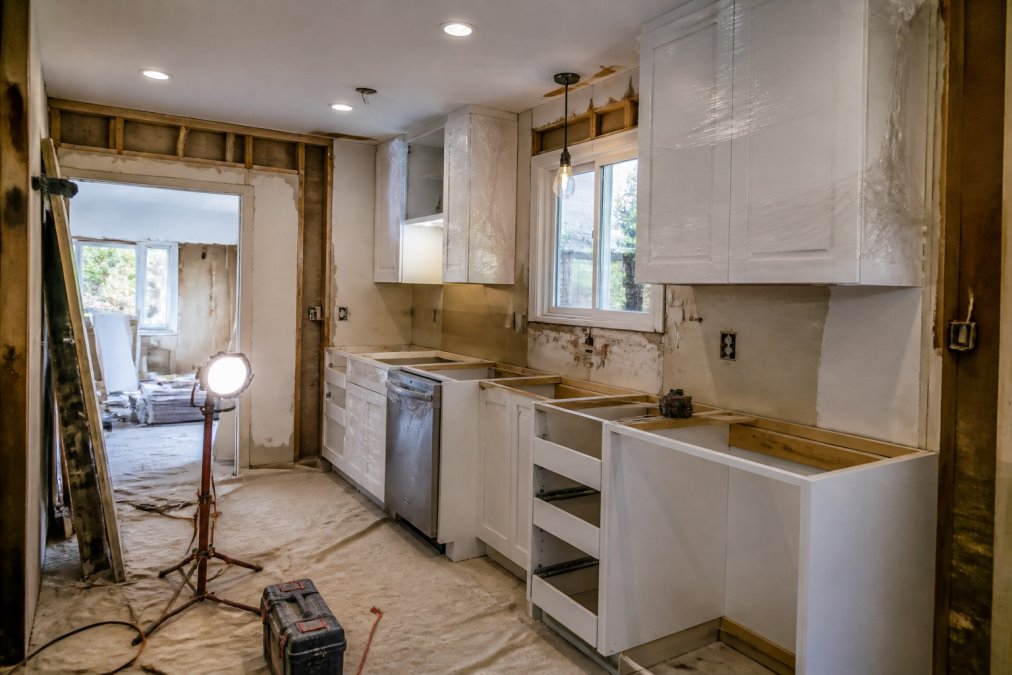 Kitchen under active renovation showing cabinetry installation in progress with temporary plastic sheeting, exposed framing, and work-in-progress state with bright work lights