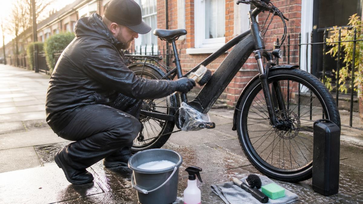 A commuter in waterproof clothing cleaning an e-bike frame with a soft brush and bucket of soapy water outside a terraced house, battery removed and motor area covered, morning light