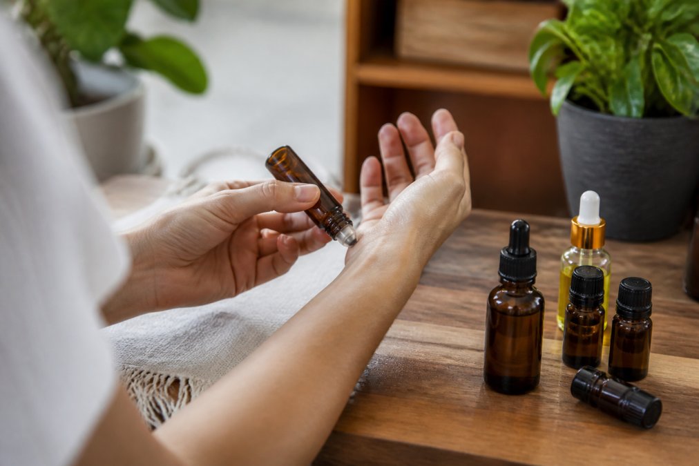 Hands applying essential oil roll-on to inner wrist pulse point, with amber glass bottles of oils and jojoba oil visible on wooden surface