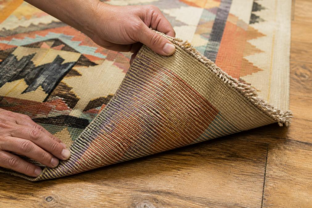 Close-up of hands flipping over a flatweave kilim rug on a wooden floor, showing the reverse side with visible weft threads and knotted fringe at the edge