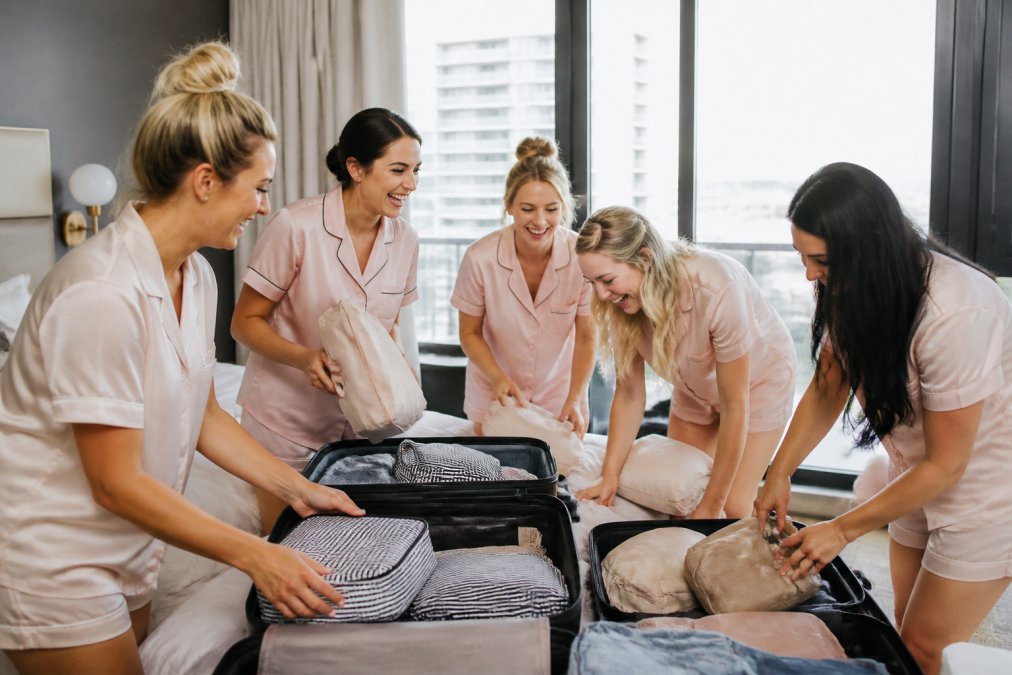 A joyful group of bridesmaids in matching casual outfits laughing while packing suitcases in a bright, modern hotel room with natural light streaming through large windows