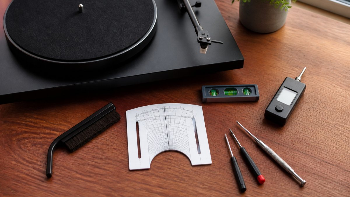 Overhead close-up of seven turntable setup tools laid out on a wooden desk beside a record player, including a stylus force gauge, alignment protractor, spirit level, cleaning brush and small screwdrivers, soft natural window light