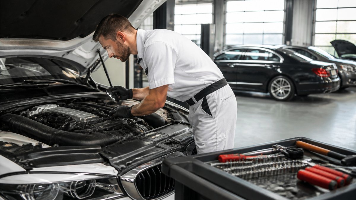 Professional ASE-certified technician in white uniform examining a European luxury vehicle engine with diagnostic equipment and tools visible in a modern, well-lit automotive shop bay