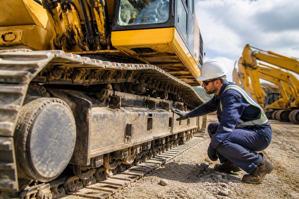 Professional technician inspecting the undercarriage and hydraulic systems of heavy construction equipment in a service yard with bright natural lighting