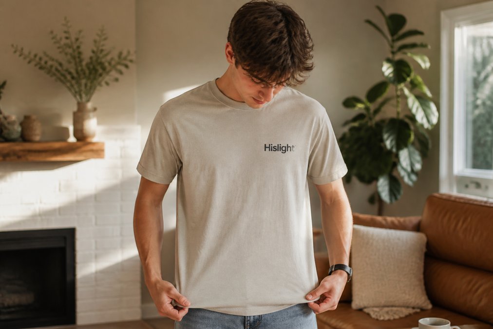 A young adult in a neutral-toned graphic tee with a subtle faith-based design, standing in a sunlit modern living room, looking down at the print with a slight smile