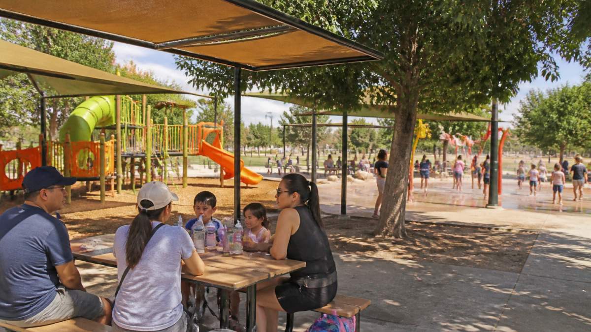 Children playing on colorful playground equipment with splash pad visible in background, families sitting at picnic tables under shade structures on a sunny afternoon