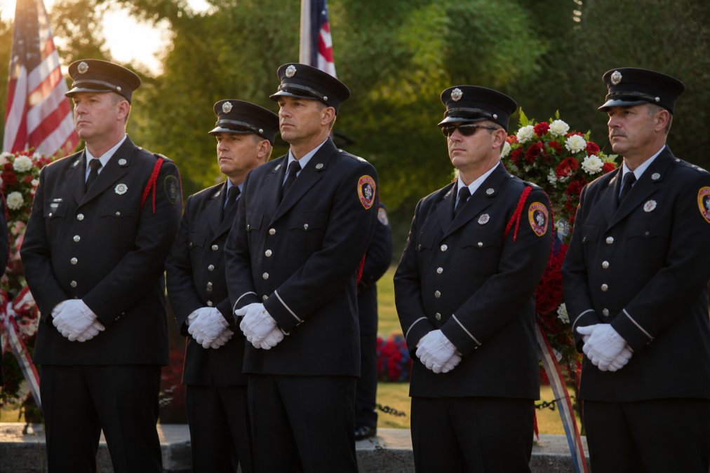 Firefighters in full dress uniforms standing together at a solemn memorial service, with wreaths and American flags visible in soft golden afternoon light