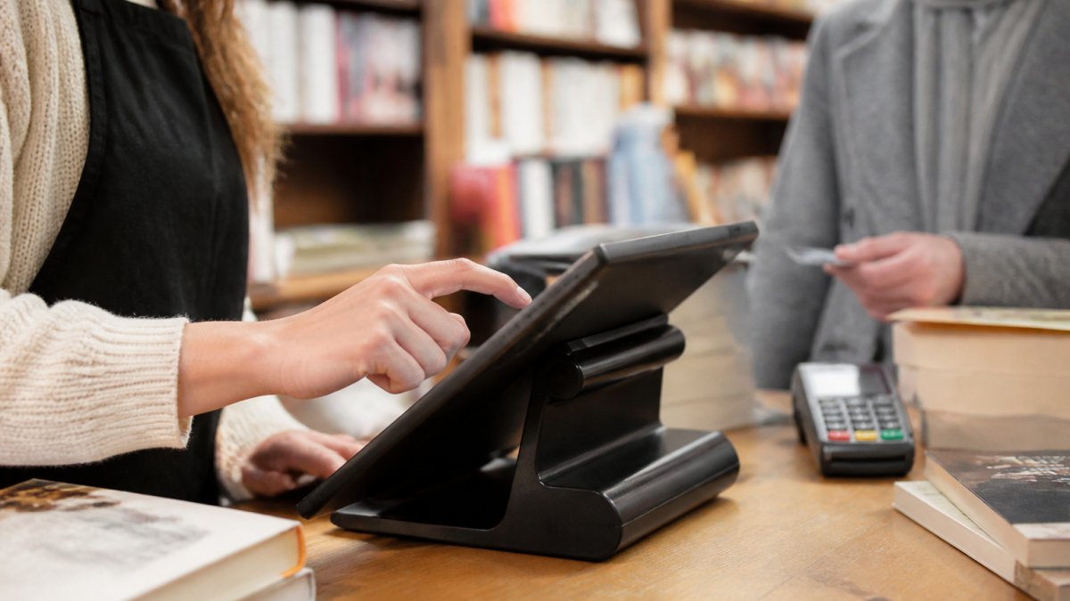 Close-up of a bookseller's hands using a touchscreen POS system at a bookstore counter, with a customer paying by card in the background