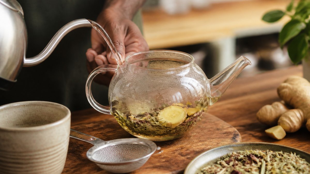 Steaming water pouring from a kettle into a glass teapot filled with dried herbs and ginger slices on a wooden counter