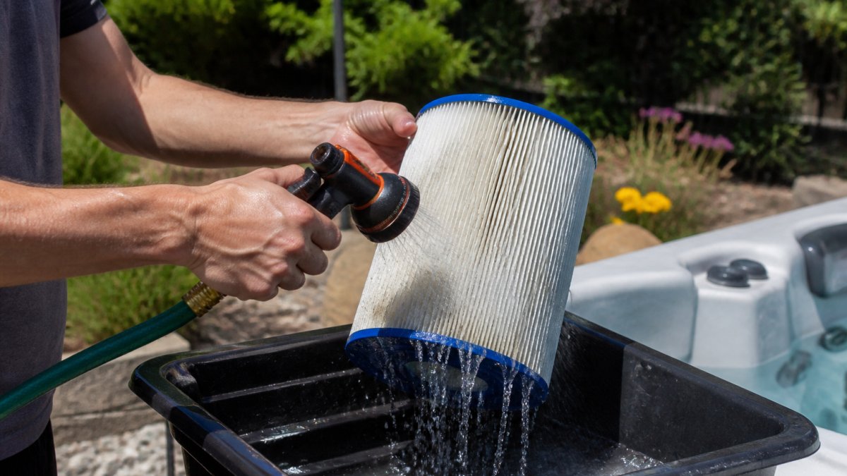 Hands rinsing a pleated hot tub filter with a garden hose nozzle in a sunny backyard
