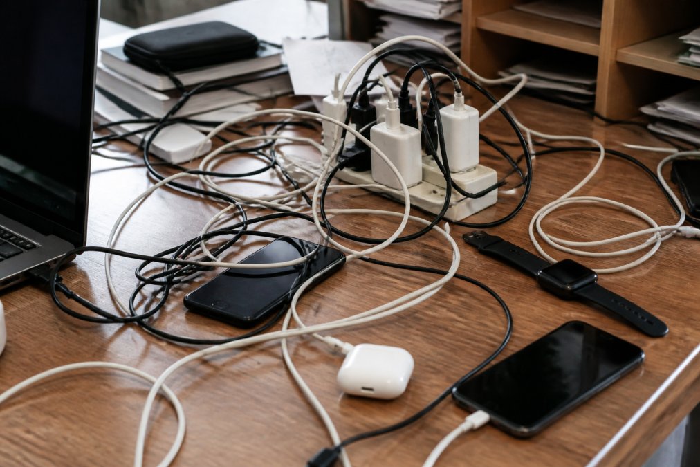 Cluttered desk with tangled cables, multiple power adapters, scattered phones and devices, cables draped across wooden surface with poor organization