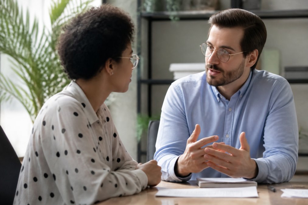 New manager having a focused one-on-one conversation with a team member at a desk, both leaning forward with engaged body language and active listening, natural office lighting