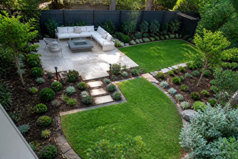 Aerial view of a suburban backyard with defined paved entertaining area, minimal lawn sections, and mulched garden beds with hardy shrubs and low-growing ground cover