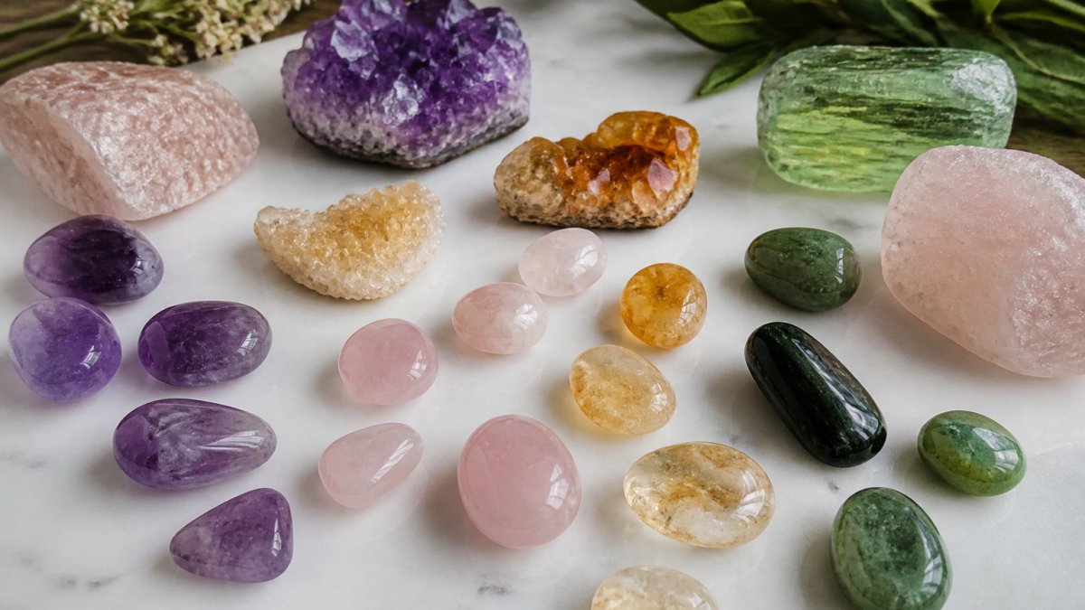An assortment of polished and tumbled healing crystals including purple amethyst, pink rose quartz, golden citrine, and green tourmaline arranged on a white surface in natural light