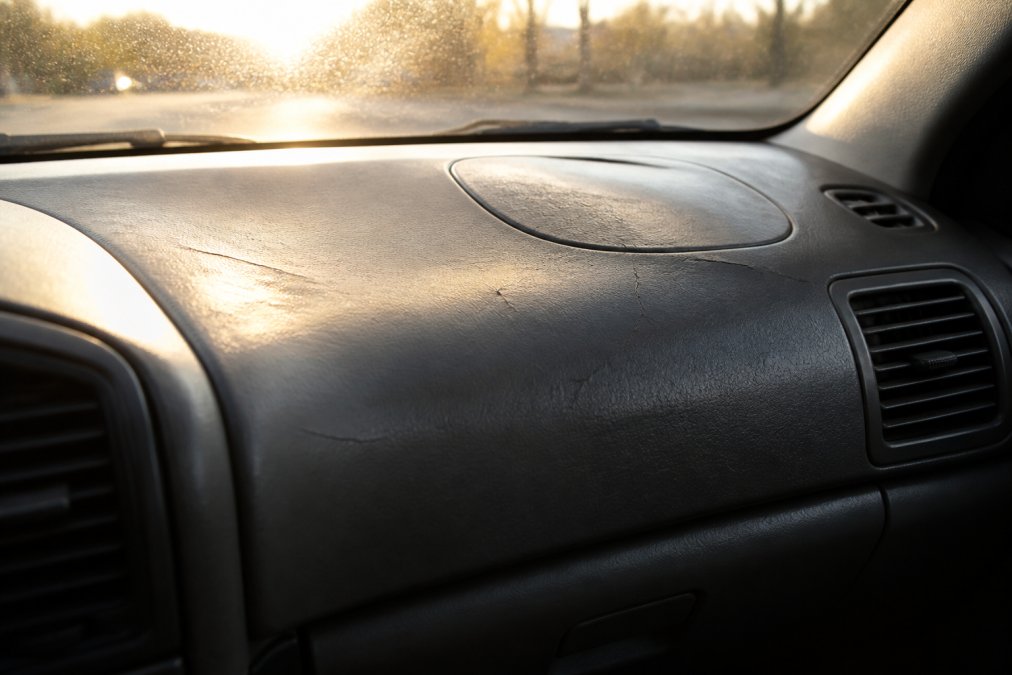 Close-up van een zonnig auto-interieur met zichtbare verkleuring en haarscheurtjes in het dashboardkunststof, fel zonlicht valt door de voorruit naar binnen
