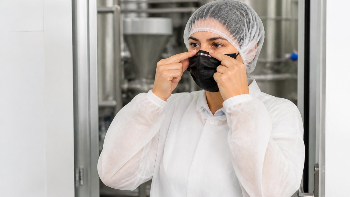 A food production worker in a hairnet, smock, and black face mask adjusting the nose wire with clean hands at the entrance to a processing area, stainless steel equipment visible behind